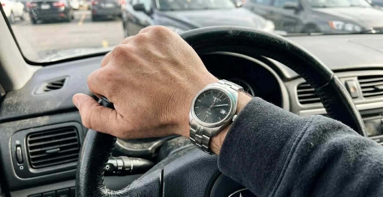 Une main sur un volant de voiture avec une montre visible au poignet, intérieur de véhicule flou en arrière-plan