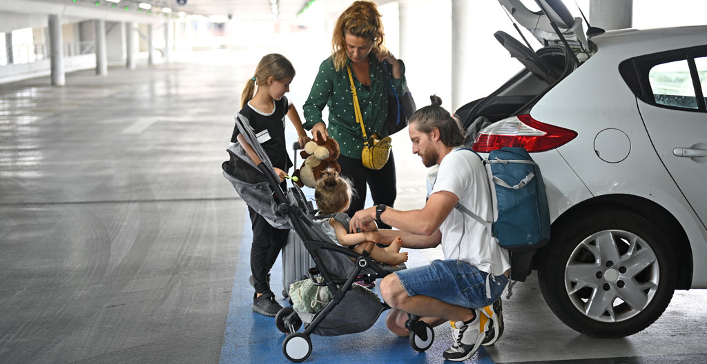 Une famille avec deux enfants et des valises debout près d'un minivan blanc sur un parking, cadrage naturel sans pose